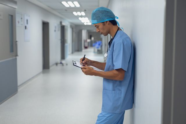 Medical professional wearing blue scrubs and a stethoscope writing on a clipboard in a hospital hallway. Useful for healthcare articles, medical service promotion, hospital advertisements, professional medical website development, or educational materials related to healthcare professions.