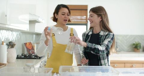 Diverse friends sorting recyclables in sunlit kitchen, pouring water and glassware at home
