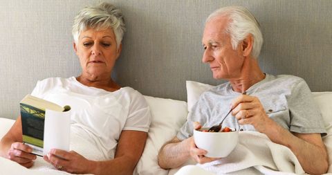 Senior Couple Relaxing in Bed Reading and Eating Breakfast