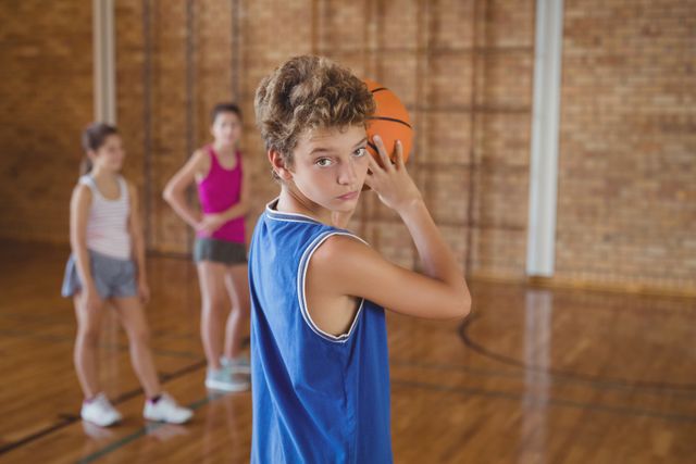 Teen boy in blue jersey preparing to take a basketball shot in a school gym, with two teammates in the background. Ideal for use in educational materials, sports training guides, youth sports promotions, and articles about physical education and teamwork.