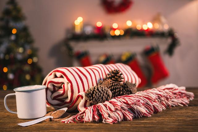 Cozy holiday scene featuring a rolled striped blanket, pine cones, and a white coffee mug on a wooden table. Background includes a decorated fireplace with stockings and candles, and a Christmas tree with lights. Ideal for use in holiday-themed promotions, home decor inspiration, and festive greeting cards.