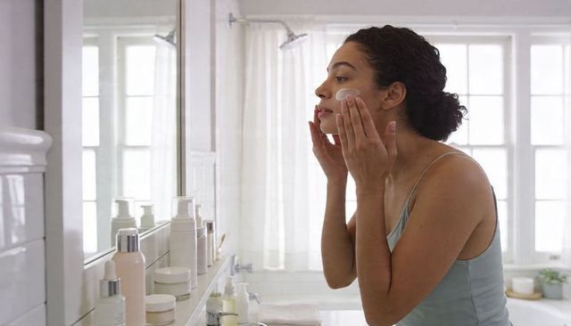 Woman applying moisturizer at vanity morning skincare routine in bright bathroom mirror