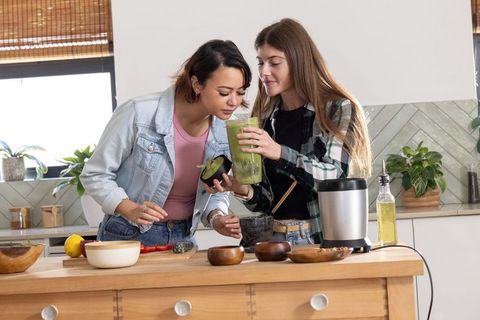 Two women preparing green smoothie at kitchen island sharing blender and smelling avocado