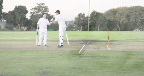Two cricketers shaking hands on cricket pitch wearing white pads holding wooden bats