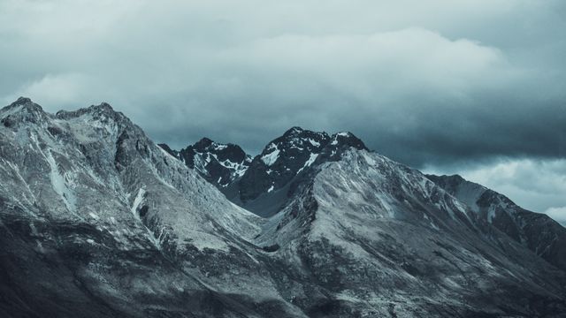 This image showcases a dramatic, snow-capped mountain range under a cloudy sky, evoking a sense of wilderness and adventure. Ideal for use in travel brochures, nature-themed projects, advertisements promoting outdoor activities, and backgrounds for website designs featuring rugged landscapes.