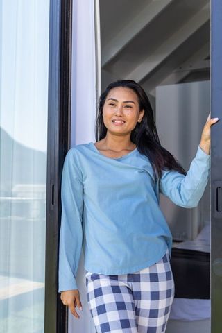 Asian woman standing at sliding door on balcony wearing blue loungewear and checkered pants