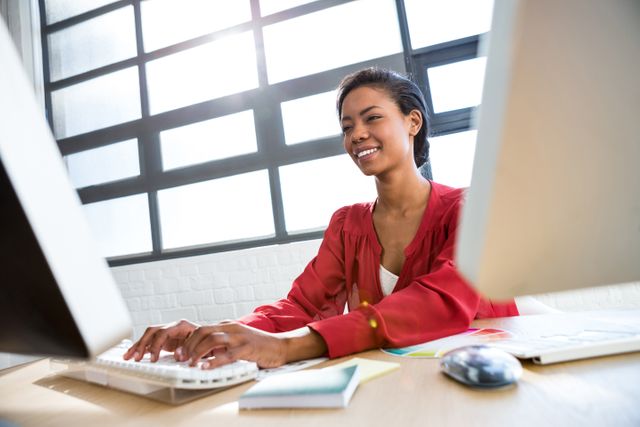 This image depicts a smiling woman working on a computer in a modern office environment. Ideal for use in business, technology, and professional settings, it can be used to illustrate concepts related to office work, productivity, and corporate culture. Suitable for websites, presentations, and marketing materials focusing on professional services, career development, and workplace environments.