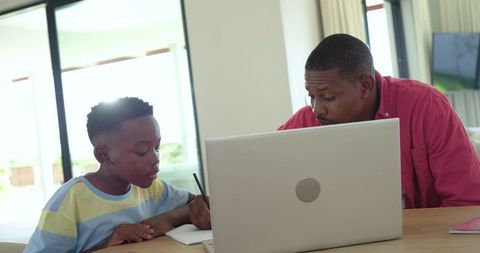 African American father guiding son with homework using laptop at daylight home study table