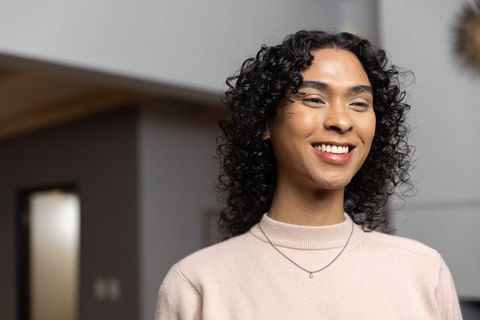 Smiling non-binary person wearing knit sweater and gold pendant in warm home interior