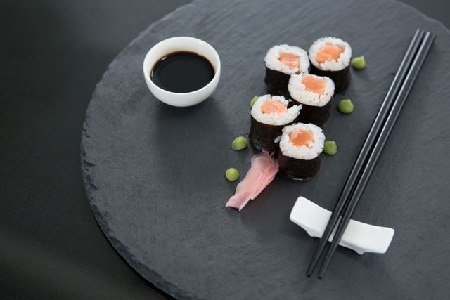 Sushi rolls with salmon and rice placed on a black slate plate, accompanied by a small bowl of soy sauce, chopsticks, pickled ginger, and wasabi. Ideal for use in articles or blogs about Japanese cuisine, sushi recipes, food photography, or restaurant advertisements.