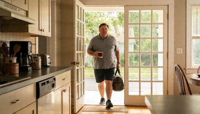 Man walking into sunlit kitchen with coffee mug and duffel bag through french doors