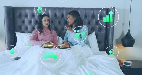 Two women sharing breakfast in bed while discussing smart home energy and finance