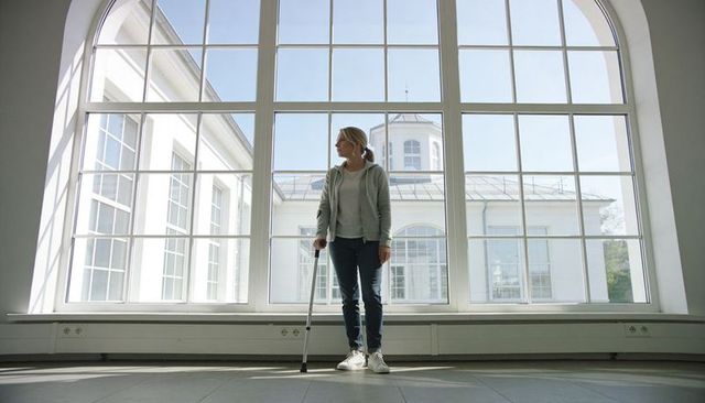 Woman using forearm crutch in bright modern interior by large arched window, daylight