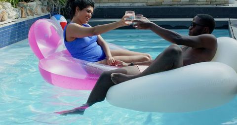 Diverse couple clinking glasses relaxing on pink and white pool inflatables for summer