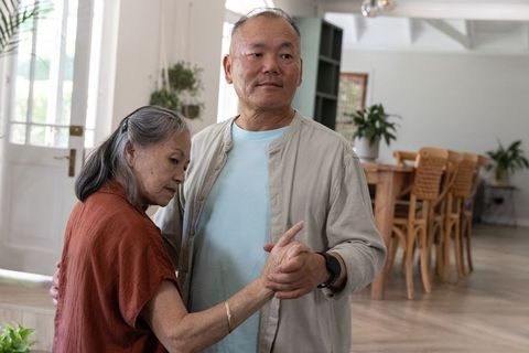 Asian senior couple swaying together at home holding hands, intimate cozy companionship