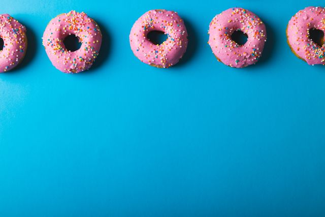 This image shows five pink donuts with colored sprinkles arranged in a row on a blue background. Perfect for use in advertisements for donut shops or bakeries, food blogs, social media posts about sweets and desserts, or promotional material for events involving treats and indulgence. The bright colors make it eye-catching for food-related publications and marketing campaigns.