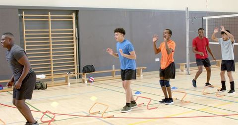 Diverse young men doing volleyball agility drills in indoor gym with hurdles and cones