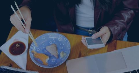 Asian woman multitasking with smartphone and chopsticks at wooden cafe table, laptop