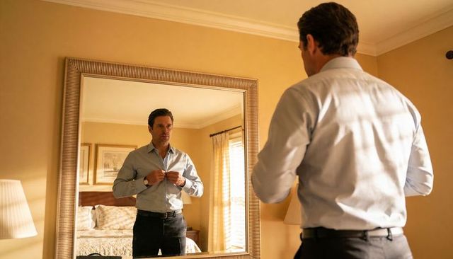 Man buttoning light blue dress shirt in bedroom mirror preparing for formal day