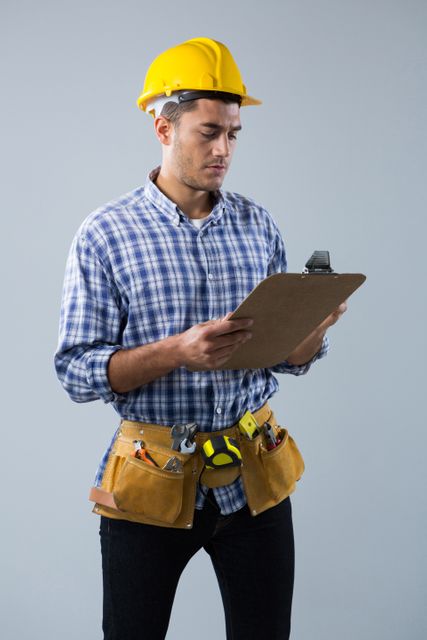 Male architect wearing hard hat and tool belt, looking at clipboard. Ideal for use in construction, engineering, and project management contexts. Suitable for illustrating concepts of planning, safety, and professional work in the building industry.