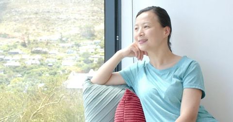 Woman relaxing on sofa by window, gazing at sunny hillside, mindful home wellbeing