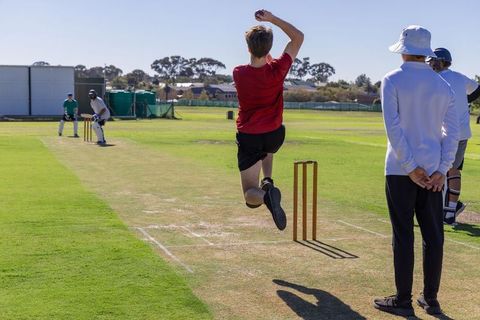Mid-air fast bowler delivering ball on sunny cricket pitch with batsman and umpire