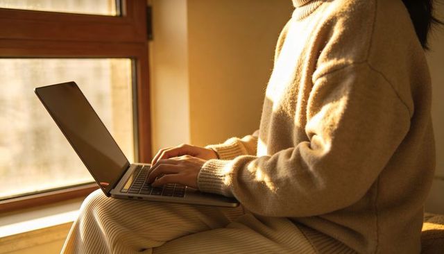 Young woman typing on laptop at sunlit window bench wearing cozy knit sweater for remote work
