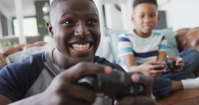 A father and his son enjoying playing video games together in a living room setting. The father is smiling broadly as he focuses on the game, while his son who's in the background, also holds a controller. This image can be used for family-related articles, parenting blogs, advertisements for gaming consoles, or general lifestyle content showcasing family bonding moments.