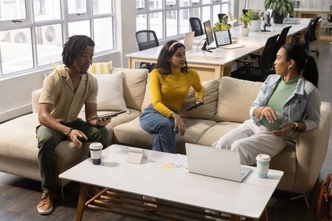 Diverse startup team collaborating on couch in modern open-plan office lounge with devices