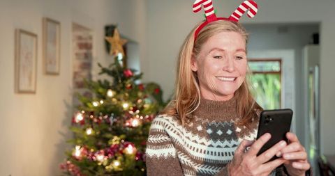 Senior woman in festive sweater using phone near christmas tree at home