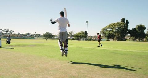 Batsman jumping and swinging bat on neighborhood cricket field with outfielders nearby