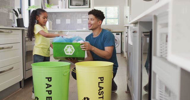 Father and young daughter are sorting recyclables together in a modern kitchen. They are placing items into appropriately labeled bins for paper and plastic. This could be used to illustrate topics like sustainable living, family bonding, and environmental education.