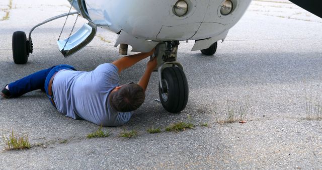 Mechanic lying on ground inspecting landing gear of small airplane on airport tarmac. Useful for articles on aviation safety, aircraft maintenance tutorials, or technical illustration for aviation mechanics.