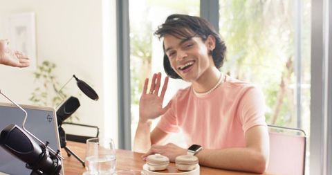 Diverse home podcast cohosts waving at camera with microphones, laptop, macarons
