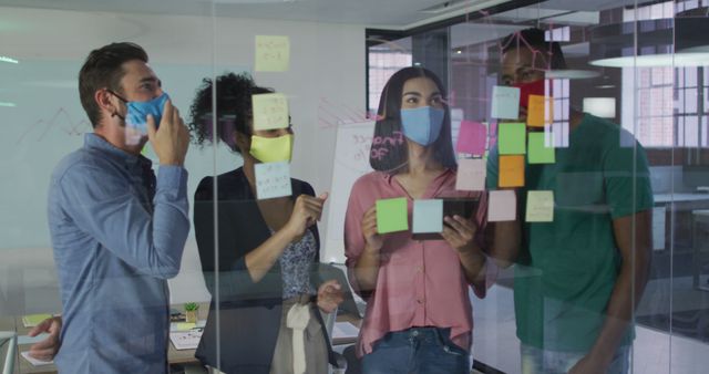 Colleagues wearing facial masks engaged in brainstorming session using sticky notes on glass wall in a modern office setting. Ideal for concepts related to teamwork, collaboration, business innovation, remote working adaptations, hybrid team interactions, and pandemic safety measures in workplaces.
