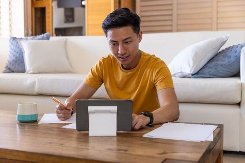 Asian man working remotely at home using tablet and taking notes, focused modern workspace