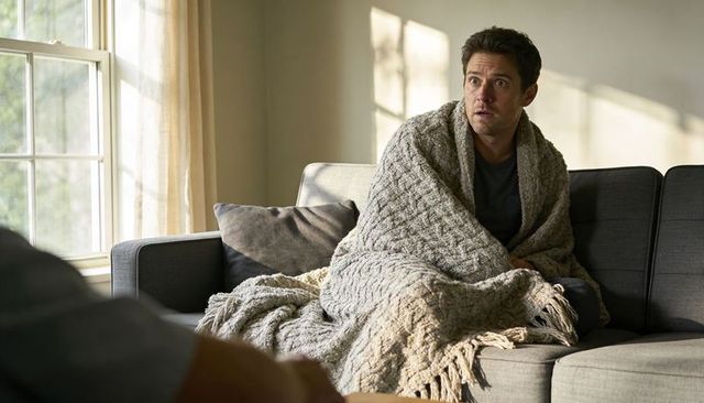 Man wrapped in chunky beige blanket sitting on modern gray sofa in sunlight looking concerned
