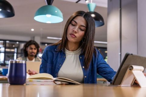Asian woman studying at shared coworking table with tablet and notebook, focused and calm