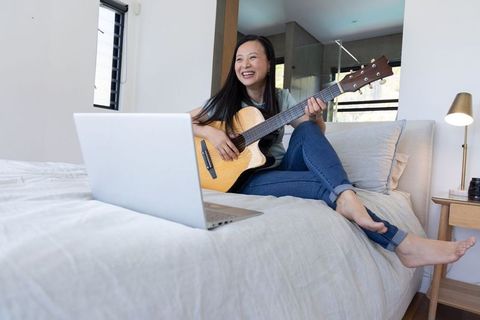 Asian woman learning acoustic guitar on bed during online lesson in modern cozy bedroom
