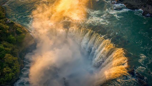 Sunlit mist rising over curved turquoise waterfall and canyon at golden hour