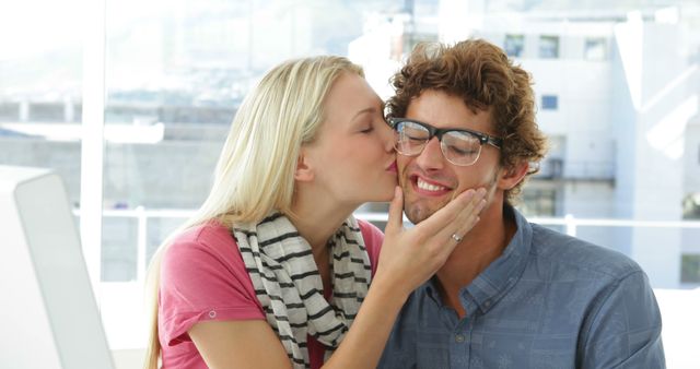 Couple showing affection by kissing in an office setting. Man wearing glasses smiles while receiving a kiss from a woman. Ideal for content related to relationships, workplace romance, lifestyle and happiness.