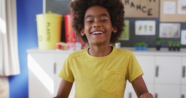 This image depicts a smiling African American boy with curly hair wearing a yellow shirt in a bright, educational classroom. He seems happy and enthusiastic about learning. Suitable for use in educational content, school brochures, back-to-school campaigns, and promotional material for children's programs.