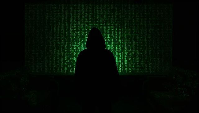 Standing hooded hacker silhouette facing cascading green code screen in dark room