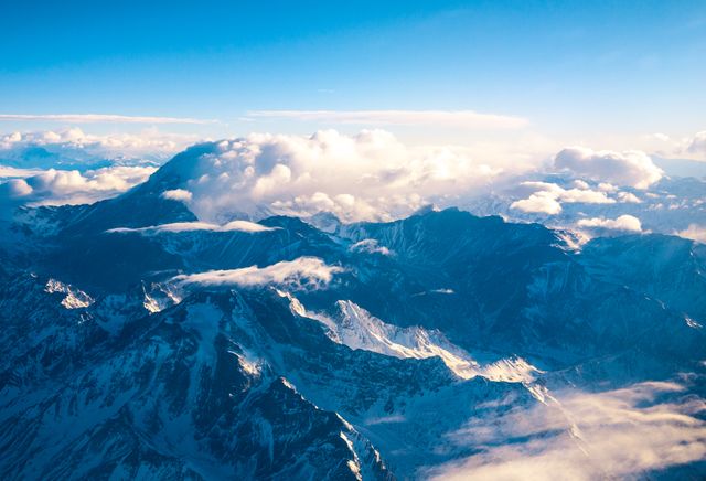 Stunning image of snow-covered mountain peaks beneath a cloudy blue sky, taken from high altitude. Ideal for nature blogs, travel websites, winter season promotions, and as a scenic background for various design projects. Captures the serene beauty and majesty of mountainous landscapes.