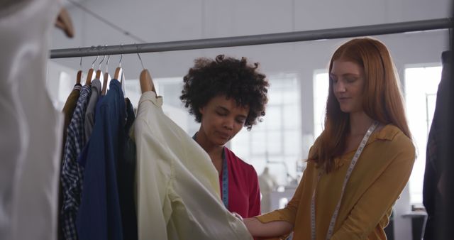 Two young fashion designers examining clothes hanging on rack in fashion studio environment. Both are focused on garment details and discussing. Measuring tapes around their necks indicate active involvement in design and sewing. Ideal for content related to fashion industry, teamwork, collaboration in creative fields, or business environments.