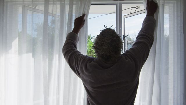 Senior African American man starts his day by opening the bedroom curtains, letting in the daylight and fresh air. This video can be used for themes related to morning routines, positive daily habits, healthy living, and promoting mental wellness. It also captures the essence of quarantine and self-isolation, relevant for health and well-being topics during the pandemic.