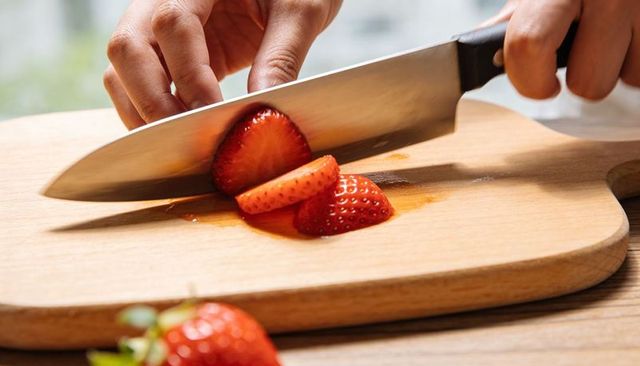 Hands slicing fresh strawberries with chef knife on wooden cutting board closeup