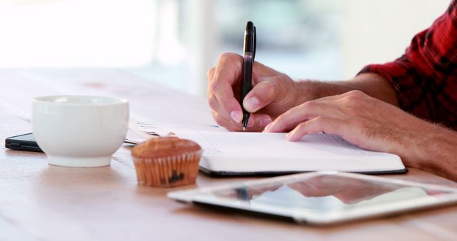 A Caucasian man is writing in a notebook at a table with a coffee cup, a muffin, and a tablet, with copy space. His focus and the presence of breakfast items suggest a productive start to his day.