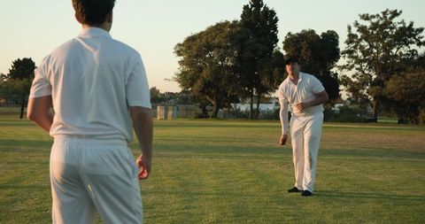 Cricket teammates practicing bowling and fielding on sunlit mown grass wearing white uniforms