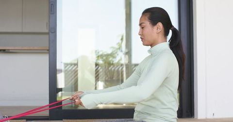Mid-adult asian woman exercising with resistance bands on patio by glass door in daylight